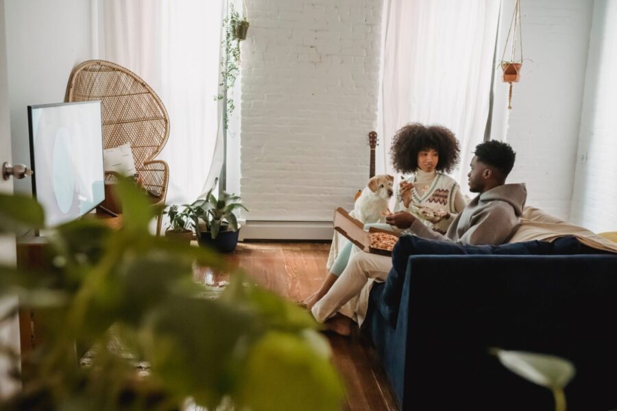 Young African American couple having snack with pizza in lounge with cozy sofa and funny white dog